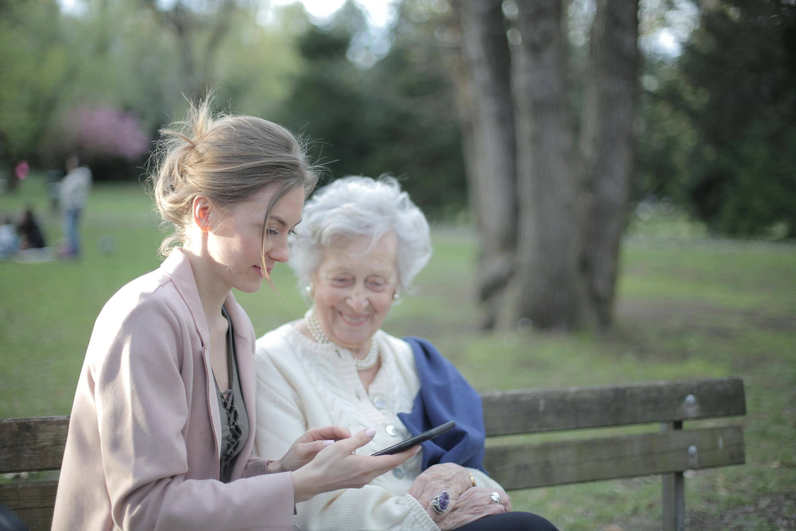 Side view of smiling adult female helping aged mom in using of mobile phone while sitting together in park